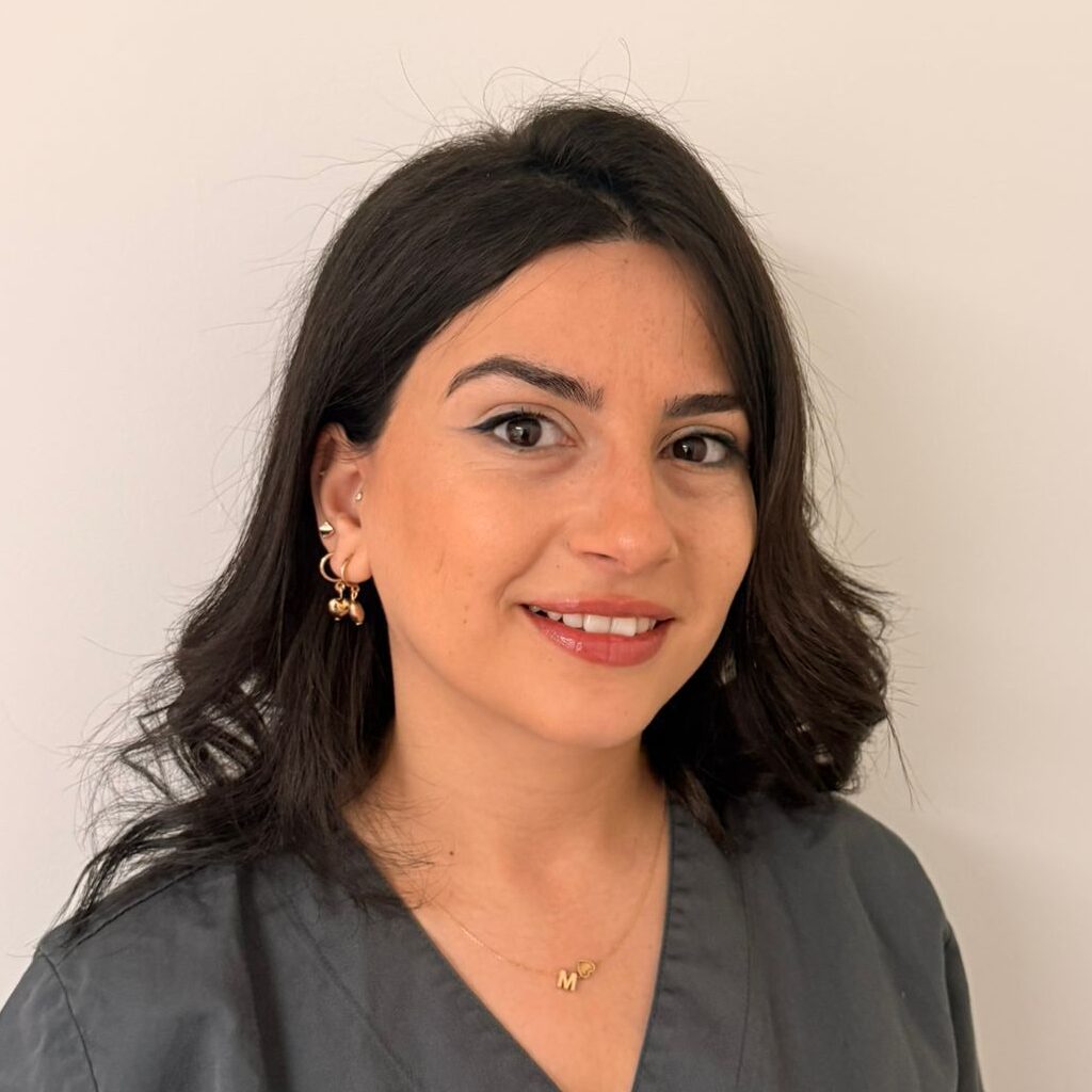 Eine lächelnde Frau mit dunklen Haaren trägt graue Kleidung vor weißer Wand